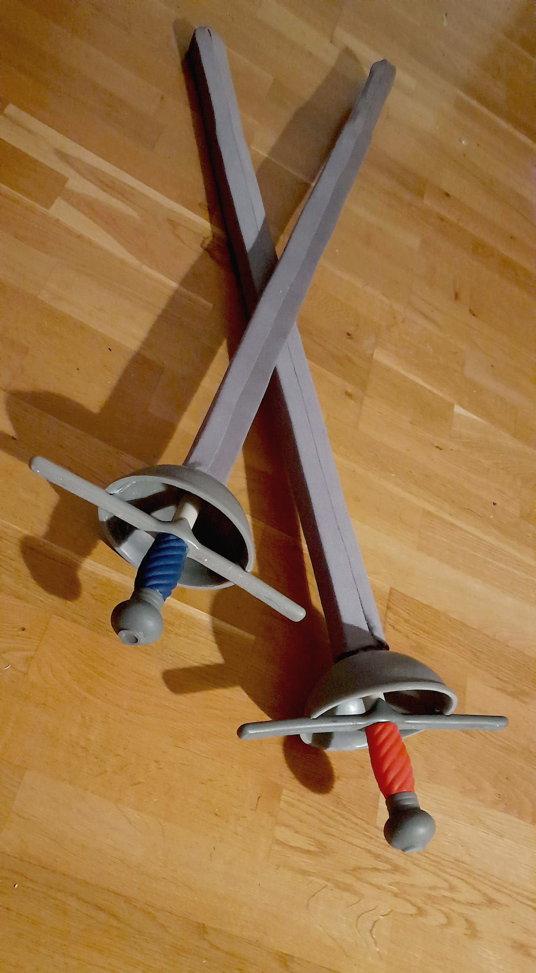 Picture of Fomp! rapiers with blue and red handles lying across each others' blades on a wooden floor.